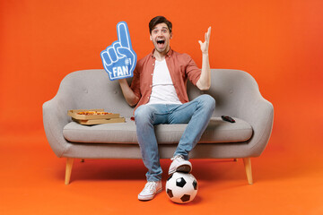 Young surprise man football fan in shirt support team with soccer ball sit on home sofa watch tv...