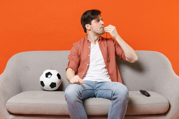 Young man football fan wearing shirt support team with soccer ball sit on sofa at home watching tv...