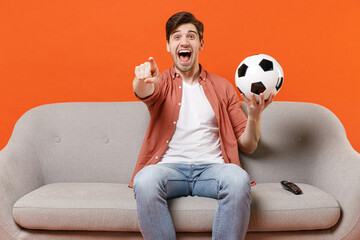 Young fun man football fan in shirt support team with soccer ball sit on sofa home watch tv live stream point index finger camera on you isolated on orange background People lifestyle sport concept