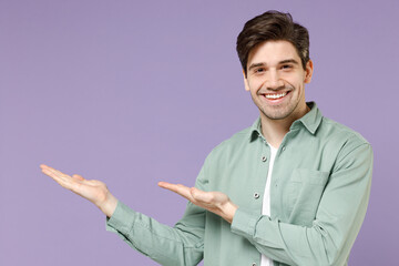 Young fun excited happy european man 20s in casual mint shirt white t-shirt point hands arms aside on workspace area mock up isolated on purple background studio portrait. People lifestyle concept