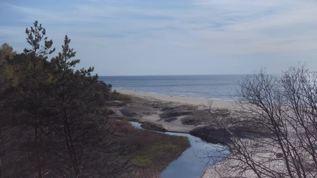 The river flows into the Baltic Sea at Saulkrasti. People walk along the yellow sand of the beach on a sunny warm spring day. Trees grow on a tall white dune in Latvia.
