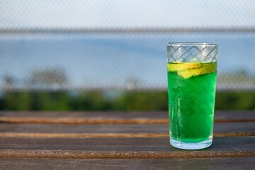 Close up wet glass of green cold mint drink, lemon slice, on wooden table, blurred blue sea background. Cooling summer drink. Summer fresh and cooling inspiration, contrast color combination. Relax.