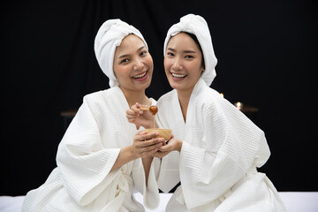 Two Asian beautiful women wearing bathrobe, towel on head, smiling, looking at camera, holding cup of honey for face, body treatment, spa and massage with dark background and candles. Beauty Concept.