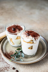 Traditional Italian dessert tiramisu in glass, light background. Coffee flavored portion dessert made of ladyfingers and mascarpone decorated with cocoa powder.
