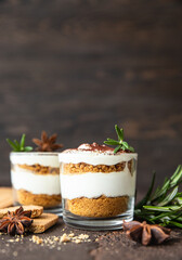 Layered dessert in glass jar with cookie crumble and whipped cream decorated with rosemary and anise, dark brown background. No bake cheesecake, trifle or pudding.