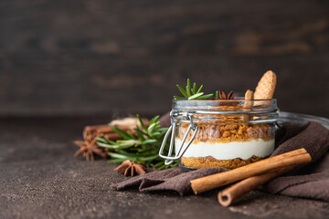 Layered dessert in glass jar with cookie crumble and whipped cream decorated with rosemary and anise, dark brown background. No bake cheesecake, trifle or pudding.