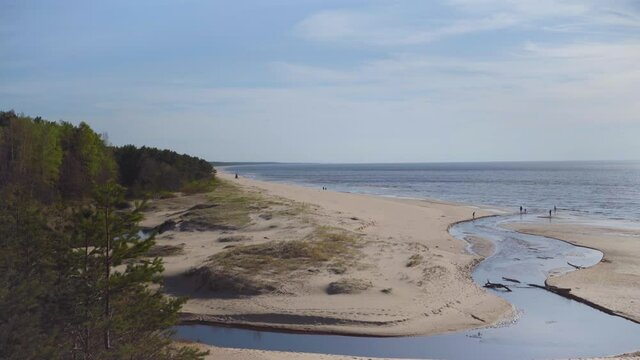 The river flows into the Baltic Sea at Saulkrasti. People walk along the yellow sand of the beach on a sunny warm spring day. Trees grow on a tall white dune in Latvia.