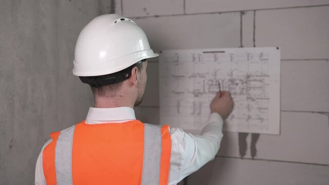 Business engineering in the premises for the construction of a high-rise building. A builder in a helmet works with blueprints.