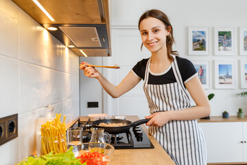 Young woman in apron cooking healthy food at modern home kitchen. Preparing meal with frying pan on gas stove. Concept of domestic lifestyle, happy housewife leisure and culinary hobby - obrazy, fototapety, plakaty
