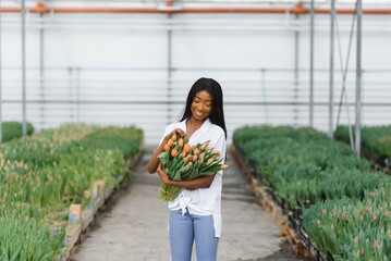 Beautiful young smiling african american girl, worker with flowers in greenhouse. Concept work in the greenhouse, flowers.