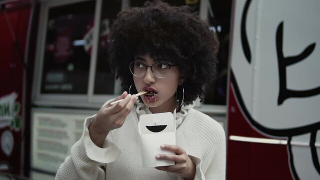 Portrait of beautiful mixed race young girl eats street food with chopsticks from white cardboard box with. Woman with afro eating outdoors