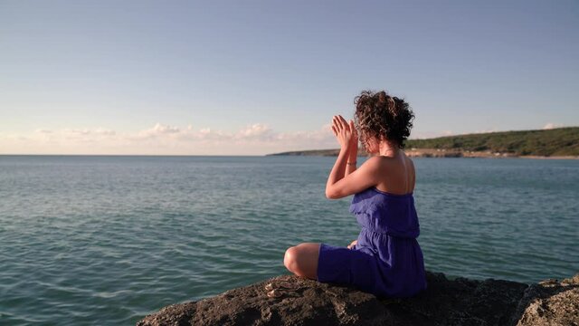 Woman in blue dress sitting on the cliff near the sea at sunset, on a windy day, doing yoga and meditation in Turkey Sinop. She is sitting in lotus pose, raising hands up then lowering them down