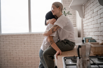 tattooed woman in glasses sitting on kitchen table and hugging brunette girlfriend