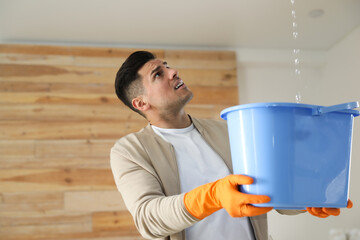 Emotional man collecting water leaking from ceiling indoors. Damaged roof