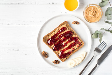Homemade peanut butter sandwich with raspberry jam and banana on white wooden background. Top view.