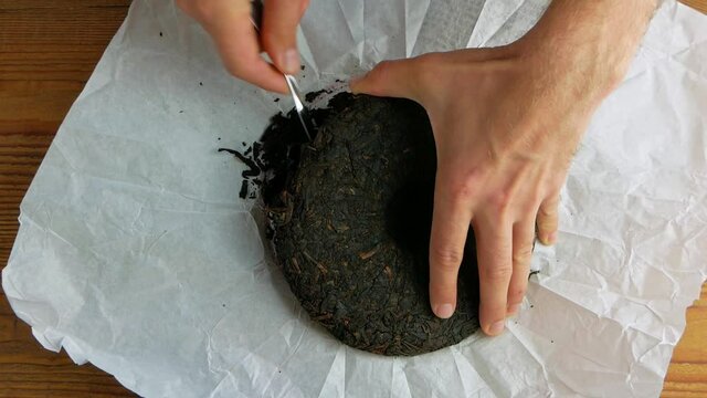 Shu Puerh cake chinese fermented black tea in white paper with knife. Male hands hold chip break pressed old round form ripened dark pu-erh into pieces. Healthy invigorating drinks coffee substitute.