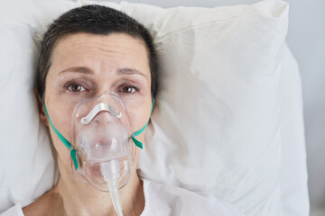 Close-up of senior woman in oxygen mask looking at camera while lying on bed on reanimation
