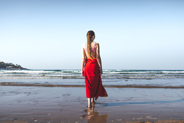 Young woman wearing a red towel and a pink bikini walking at the water edge on the beach