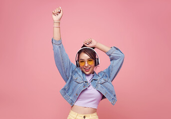 Happy beautiful Asian teen woman in yellow glasses listening music in headphones and dance on pink...