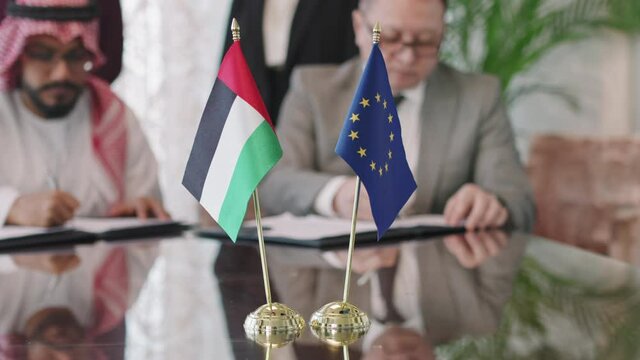 Close-up of UAE and European Union flags on negotiation table in selective focus while political representatives of these countries signing cooperation agreement in blurred background