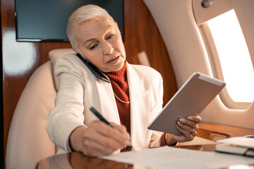 Businesswoman with digital tablet writing on paper and talking on smartphone in private plane