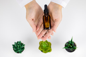 Close-up hands hold glass bottle of cosmetic oil on white background with plants. Copy space. Dermatology, healthcare, treatment, microbiome skincare, plant-based products, beauty trends concept