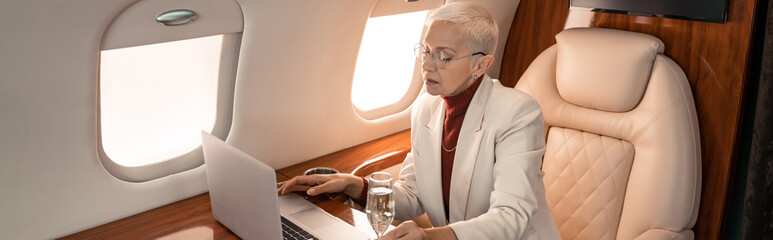 Businesswoman using laptop near glass of champagne in jet, banner