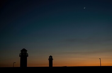 silhouette of lighthouse towers at a harbor with crescent moon in the evening