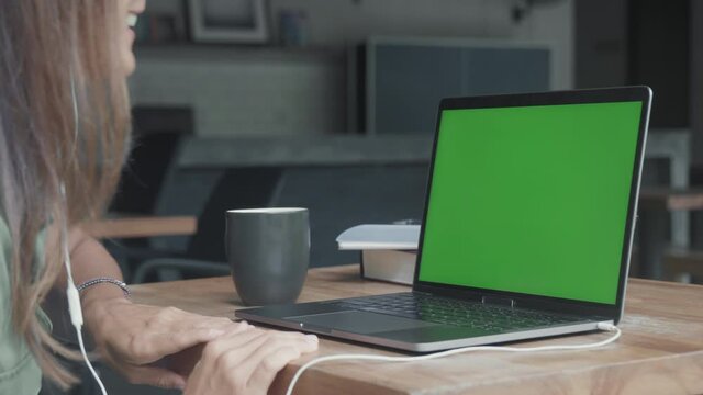 Steadicam of unrecognizable person wearing wired headphones, sitting by desk at home, talking gesticulating, drinking from mug, using laptop computer with green screen