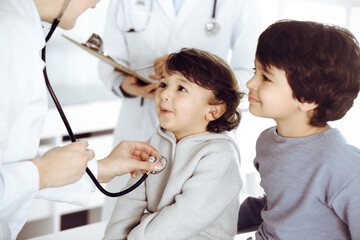 Woman-doctor examining a child patient by stethoscope. Cute arab toddler and his brother at physician appointment. Medicine concept