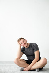 Obraz premium Positive man. Home relax. Lockdown lifestyle. Harmony peace. Handsome happy guy in t-shirt shorts sitting floor carpet in lotus pose leaning hand on leg light wall interior background copy space.