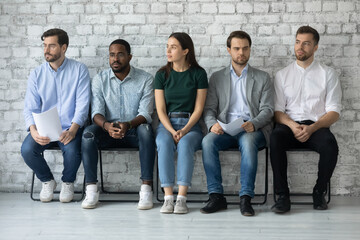 Worried unemployed professionals sitting in queue, holding paper resumes, waiting for job interview appointment with recruiter. Line of diverse candidates expecting employer decision in corridor