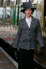Beautiful english woman dressed in 1920s costume walking on railway platform with train in...