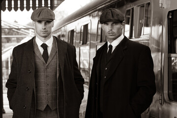 Handsome English gangsters smoking at railway station with train in the background