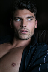 Fototapeta premium Portrait of attractive young man with stubble and open shirt revealing defined pecs, sitting in leather armchair