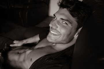 Portrait of attractive young man with beard and open shirt revealing sixpack abs, sitting in...