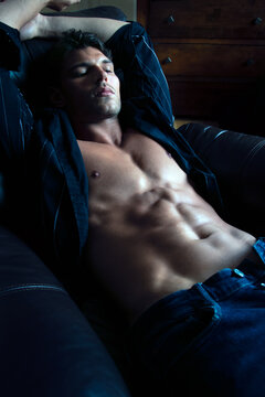 Portrait of attractive young man with stubble and open shirt revealing defined pecs, abs, lying on leather armchair