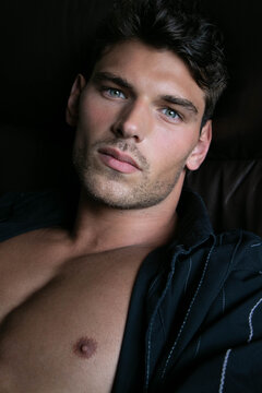 Portrait of attractive young man with stubble and open shirt revealing defined pecs, sitting in leather armchair
