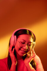 Music chill. Neon light portrait. Futuristic technology. Happy Asian girl in pink enjoying listening song in headphones isolated on orange color gradient copy space background.