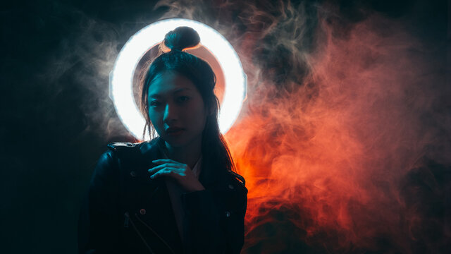Cyberpunk fashion. Night portrait. Futuristic beauty. Blue neon light Asian girl in black leather jacket with white glowing halo in red color smoke on dark copy space background.