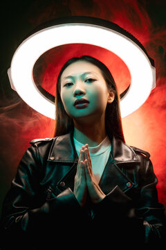 Cyberpunk People. Spiritual Portrait. Future Faith. Blue Neon Light Art Portrait Of Asian Prayer Girl In Black Leather Jacket Namaste With White Halo In Red Color Smoke On Dark.