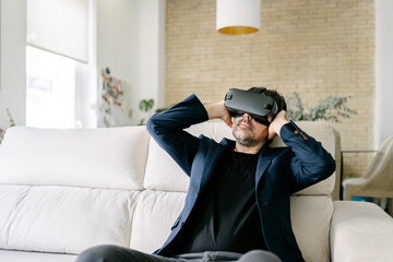 Adult man sitting on a modern sofa experiencing virtual reality in the interior of a contemporary design house. Virtual reality goggles