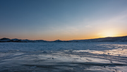Fototapeta na wymiar Sunset over the frozen lake plain. The sun is hidden behind a ridge, the sky is highlighted in orange. Reflections on ice. Baikal