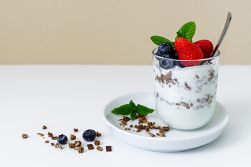 Joghurt mit Granola, Erdbeeren und Heidelbeeren in einem Glas geschichtet. Modern, weißer Tisch, beige Wand, gesundes Frühstück.
