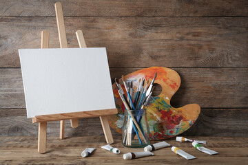 Easel with blank canvas, brushes, paints and palette on wooden table