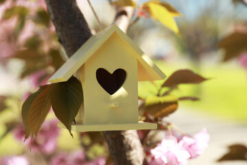 Fototapeta na wymiar Yellow bird house with heart shaped hole hanging on tree branch outdoors