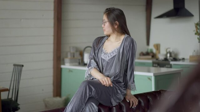 Portrait of dissatisfied young Asian woman in eyeglasses and nightie putting away business graph shaking head. Young beautiful intelligent lady having strategy problems thinking in home office