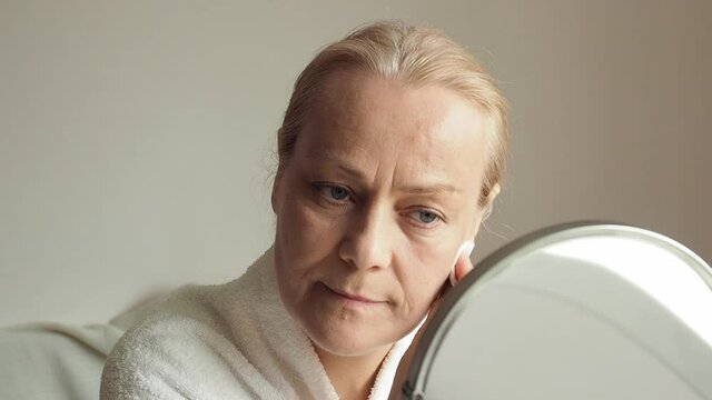 Senior woman in white bathrobe uses cotton pads to cleanse her face while looking in the mirror.