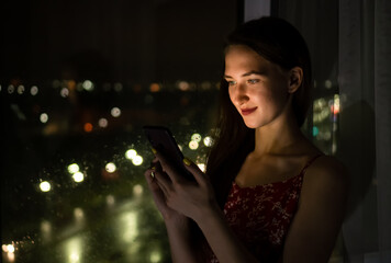 Portrait of a beautiful young woman in a red dress and with red lipstick on her lips standing near the window at night holding a mobile phone near her face, soft light from the phone falls on her face