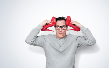 A funny and amusing man with glasses holding female red high-heeled shoes near his head in both hands, on light background.